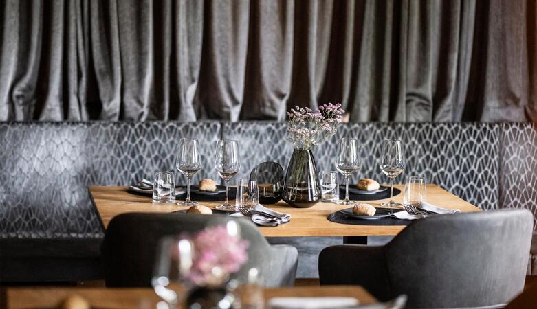 Sala da pranzo moderna con sedute rivestite in grigio scuro, tavoli in legno e una lunga tenda scura sullo sfondo; un tavolo apparecchiato per quattro persone con un vaso di fiori al centro.