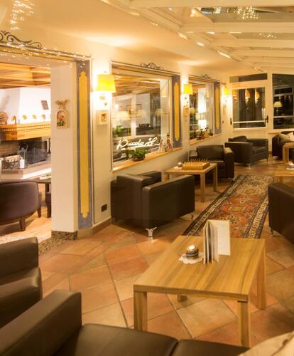 Lounge area of Hotel Piccolo with dark armchairs, wooden tables and warm lighting; on the left, the reception can be seen through a glass pane.