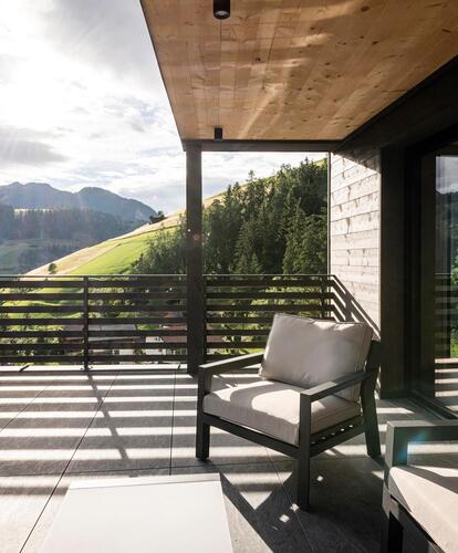 Ampia terrazza dell’hotel con sedute e facciata in vetro; sullo sfondo si vedono colline verdi, bosco e montagne illuminate dal sole.