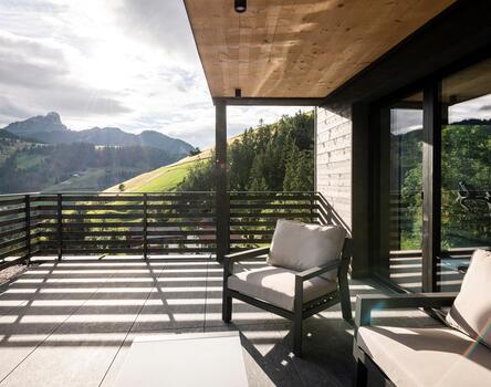 Große Hotelterrasse mit Sitzmöbeln und Glasfront; im Hintergrund sind grüne Hügel, Wald und Berge im Sonnenlicht zu sehen.