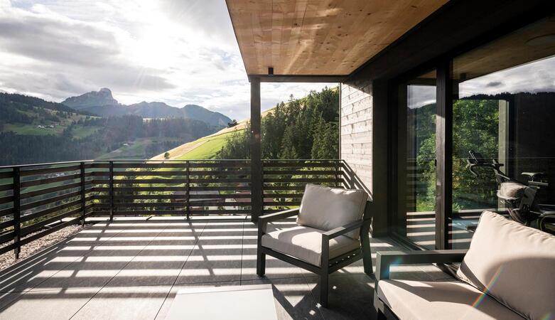 Große Hotelterrasse mit Sitzmöbeln und Glasfront; im Hintergrund sind grüne Hügel, Wald und Berge im Sonnenlicht zu sehen.
