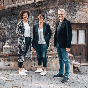 Three hosts of Hotel Pfösl stand smiling in front of an old stone wall; in the background, a wooden door and a wooden façade element can be seen.