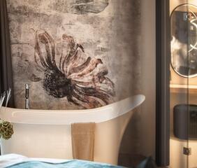 Camera d’hotel moderna con vasca da bagno freestanding davanti a una parete in pietra, motivo decorativo a parete, letto in primo piano e area bagno illuminata.