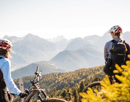 Due ciclisti su strada stanno con le loro biciclette su una collina e osservano un paesaggio montuoso alla luce calda del tramonto, con fiori gialli in primo piano