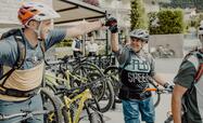 Zwei Biker in Fahrradbekleidung und Helm stehen neben ihren Mountainbikes, unterhalten sich und geben sich die Hand; im Hintergrund Häuser.