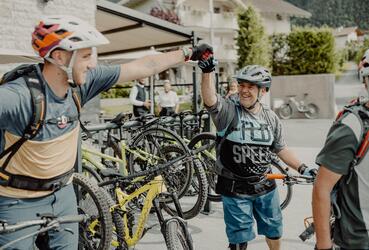 Zwei Biker in Fahrradbekleidung und Helm stehen neben ihren Mountainbikes, unterhalten sich und geben sich die Hand; im Hintergrund Häuser.