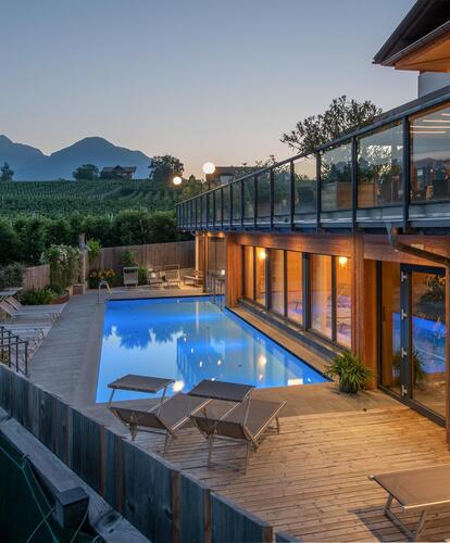 Foto serale di un hotel con area piscina illuminata, terrazza e finestre panoramiche; sullo sfondo paesaggio montano e cielo sereno.