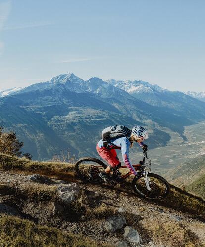 A female biker on a narrow mountain trail; the summer mountain landscape is visible in the background.