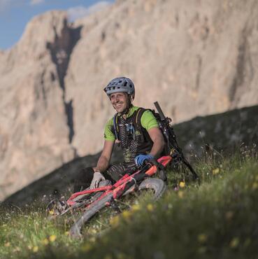 Der Gastgeber Gunther Linder sitzt neben seinem Fahrrad auf einer Wiese; sind die massiven Dolomiten zu sehen.