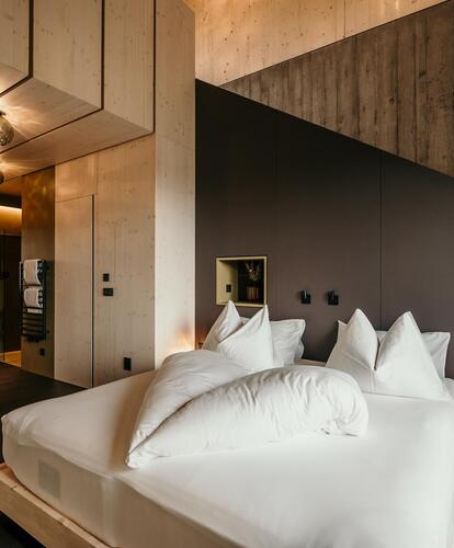 Modernes Hotelzimmer mit dunklen Wänden und Boden; eine Frau im weißen Bademantel liegt auf einem großen Bett mit weißen Kissen.