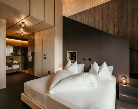 Modernes Hotelzimmer mit dunklen Wänden und Boden; eine Frau im weißen Bademantel liegt auf einem großen Bett mit weißen Kissen.