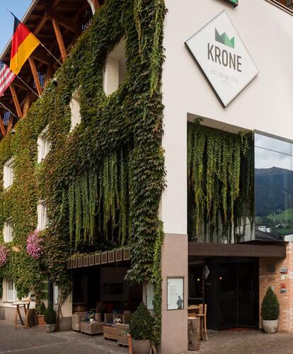 Si vede l'hotel moderno Krone a Bressanone; su una strada, facciata parzialmente ricoperta di piante verdi, diverse bandiere appese sul fronte, grande finestra di vetro, ingresso e logo visibili.