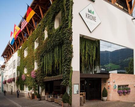 Zu sehen ist das moderne Hotel Krone in Brixen; an einer Straße, Fassade teilweise mit grünen Pflanzen bedeckt, mehrere Fahnen hängen an der Vorderseite, großes Glasfenster, Eingang und Logo sichtbar.