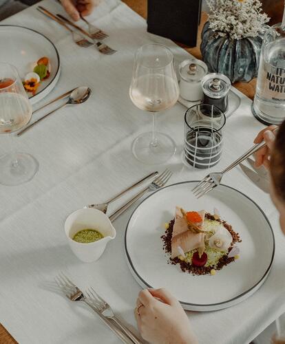 Two people are sitting at a table, with a fish dish on the plate, glasses and cutlery beside it; in the foreground, decorations with flowers.