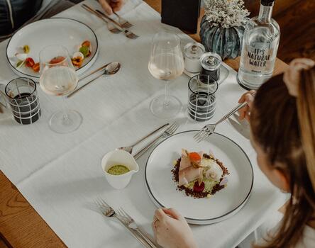 Two people are sitting at a table, with a fish dish on the plate, glasses and cutlery beside it; in the foreground, decorations with flowers.