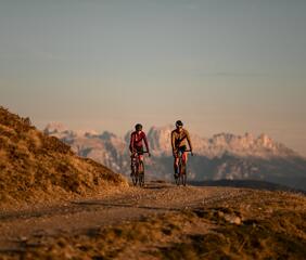 Zwei Radfahrer stehen mit ihren Fahrrädern auf einem Bergweg und blicken auf eine Berglandschaft im Abendlicht