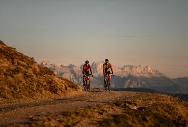 Zwei Radfahrer stehen mit ihren Fahrrädern auf einem Bergweg und blicken auf eine Berglandschaft im Abendlicht