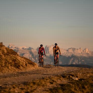 Due ciclisti con le loro biciclette stanno su un sentiero di montagna e guardano un paesaggio alpino alla luce del tramonto