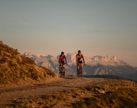 Two cyclists stand with their bikes on a mountain path, looking at an alpine landscape in the evening light