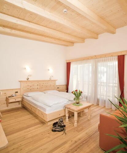 Camera d’hotel con letto matrimoniale, pavimento in legno e soffitto chiaro in legno; a destra si trova un’area salotto arancione, in primo piano si vedono una pianta e scarpe da trekking.