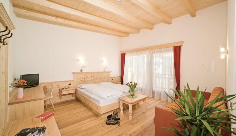 Camera d’hotel con letto matrimoniale, pavimento in legno e soffitto chiaro in legno; a destra si trova un’area salotto arancione, in primo piano si vedono una pianta e scarpe da trekking.