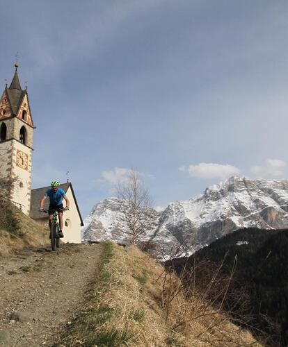 A cyclist rides downhill along a rocky path; a church, mountains and woodland can be seen in the background.