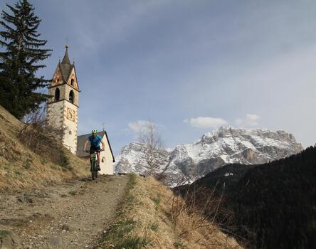 A cyclist rides downhill along a rocky path; a church, mountains and woodland can be seen in the background.