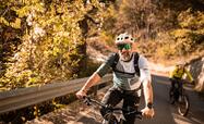 Two cyclists wearing helmets are riding up a steep road; autumn deciduous trees are visible in the background.