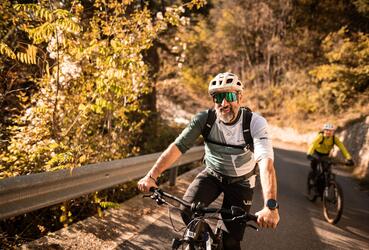 Zwei Fahrradfahrer mit Helm fahren eine steile Straße hoch; im Hintergrund sind herbstliche Laubbäume zu sehen.