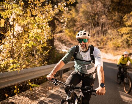 Two cyclists wearing helmets are riding up a steep road; autumn deciduous trees are visible in the background.