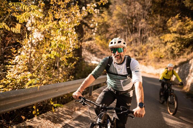 Zwei Fahrradfahrer mit Helm fahren eine steile Straße hoch; im Hintergrund sind herbstliche Laubbäume zu sehen.