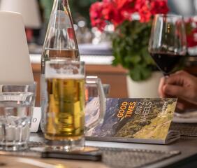 A glass of beer on a table next to a magazine, glasses, and a flower vase
