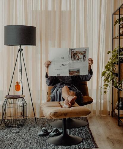 Person lying relaxed on an armchair, holding up a newspaper; in the room, a black floor lamp, a rug, and plants in the background