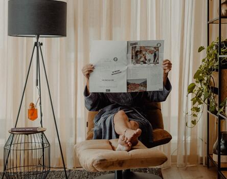 Person lying relaxed on an armchair, holding up a newspaper; in the room, a black floor lamp, a rug, and plants in the background