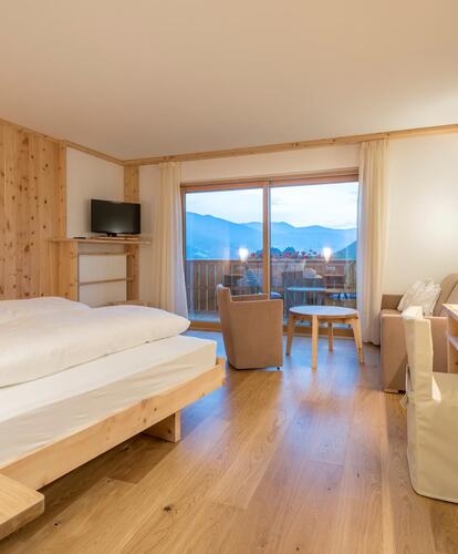 Camera d’albergo luminosa con letto matrimoniale, mobili in legno, scrivania, sedie e finestre panoramiche con vista sulle montagne; la luce del giorno entra dalle finestre.
