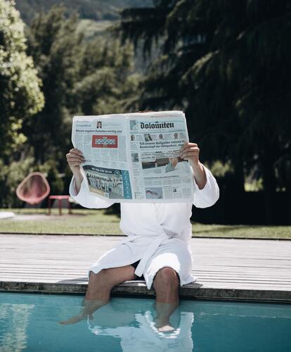 A man is sitting by the outdoor pool and reading a newspaper