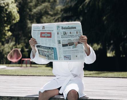 A man is sitting by the outdoor pool and reading a newspaper
